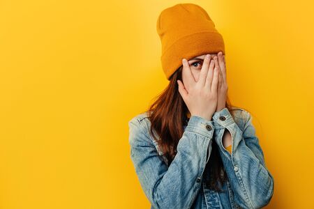 Shocked frightened young woman peeks through fingers, stares with widely opened eyes, wears denim jacket and hat, curious female student peek cover hide face in hands, isolated over yellow backgroundの写真素材