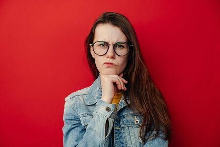 Young woman in glasses curves lips and frowns eyebrows, keeps hand under chin, looks with discontent expression, wears denim jacket, stands over red studio wall. Human facial expressions and feelingsの写真素材
