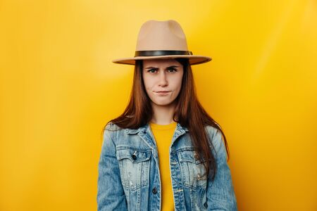 Serious unhappy woman in hat frowns from displeasure, raises eyebrows, dissatisfied with something, wears denim jacket, isolated over yellow background. Negative human emotions and feelings conceptの写真素材