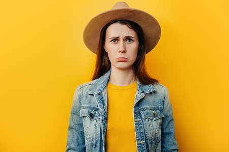 Upset young woman in hat purses lower lip, pouting and has displeased look, looks sadly at camera,...