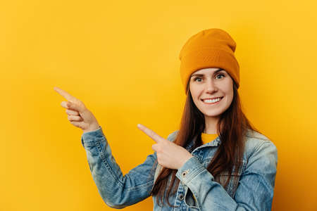 Cheerful girl in hat looking camera pointing showing fingers hands on workspace copy space mockup template. Close up of young smiling woman isolated on yellow studio background, wears denim jacket.の写真素材
