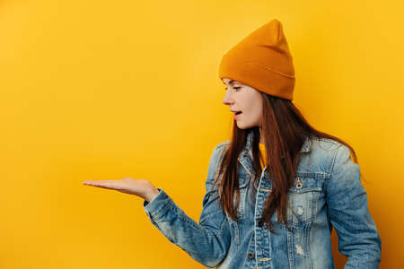 Smiling young woman in hat looks pleasant to her palm hand, showing something in her hand, dressed in denim jacket, isolated on yellow background with copy space. People sincere emotions conceptの写真素材