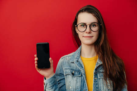 Happy young woman holds modern cell phone, shows mobile phone with empty space, satisfied with its...