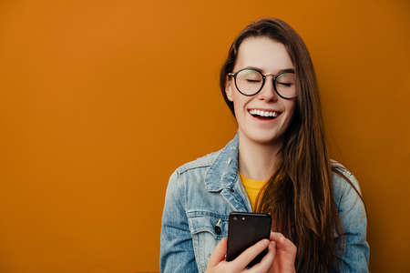 Smiling happy young woman in eyewear holds smartphone, smiles positively, creats own blog, wears...