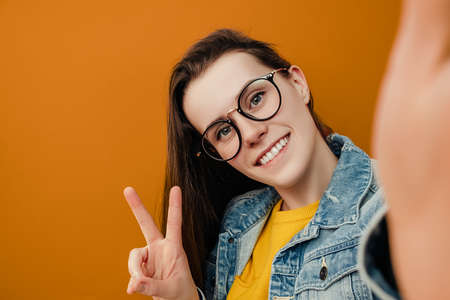 Portrait of cute brunette woman in eyewear, makes peace gesture, takes selfie on smartphone, has fun with cellular, dressed in denim jacket, poses against brown studio background with copy spaceの写真素材
