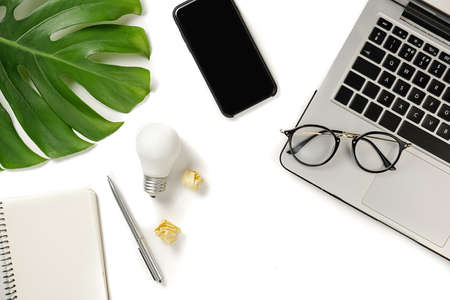 Creative top view flat lay of modern workspace with light bulb, laptop, pen, monstera leaves, eyeglasses, fresh apple, smartphone and notebook with copy space on white background minimal style.の写真素材