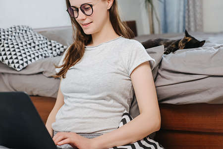 Close up of happy pregnant woman in eyeglasses sitting on floor at home working on laptop near lying cute cat, future mom searching for information on Internet. Concept of distant workの写真素材