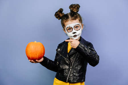 Cute preschool girl point finger at small pumpkin, wears frightening makeup, celebrates Halloween...