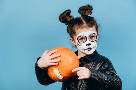 Close up portrait of zombie little girl isolated on blue studio background hold orang pumpkin, wears...