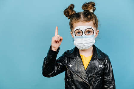 Little girl with bright scary makeup wears protective medical mask, points away, demonstrates something at blank copy space for your advertisment, isolated over on blue background. Happy Halloween!の写真素材