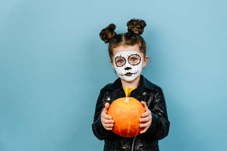 Small child hold orange pumpkin, looking at camera, dressed in black leather jacket, isolated on blue studio background. Little girl wears frightening makeup. Kid prepares for mexican day of deadの写真素材
