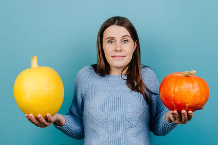 Studio shot of puzzled female shrugs shoulders, has uncertain facial expression, can't choose which pumpkin, wears sweater, models indoor over blue studio background. People and autumn conceptの写真素材