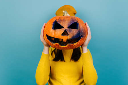 Close up portrait of young woman hiding her face behind big carved spooky pumpkin, dressed in orange...