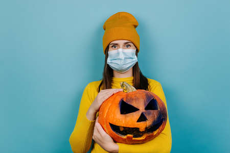 Portrait of millennial woman wears protective medical mask, holds spooky carved pumpkin, dressed in...