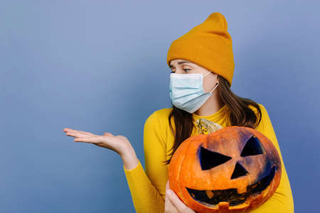 Young woman in medical mask, hold spooky pumpkin, looks on showing empty copy space on open hand...