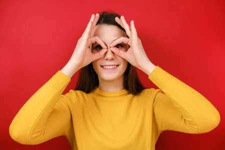 Cheerful funny young woman having fun making glasses shape with hands, does funny face with eyes...