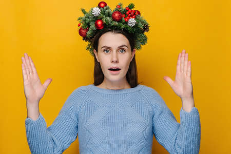 Surprised young woman showing big size gesture with hands, impressed girl demonstrate something wide, wears handmade wreath and blue sweater, isolated on yellow studio wall. Merry christmas conceptの写真素材