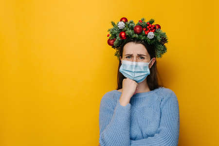 Unhappy young female wearing medical face mask, looks sad, shrugs with crossed arms, wears spruce holiday wreath around head and winter sweater, isolated on yellow studio wall with copy spaceの写真素材