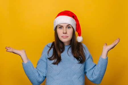 Doubting young girl shrug shoulders, looks, confused and looking to hand palms, wears Christmas hat and blue sweater, isolated on yellow studio wall. New Year celebration merry holidayの写真素材
