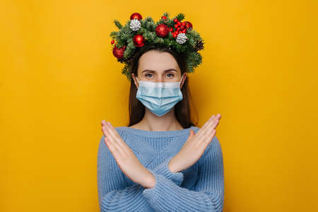 Dissatisfied young woman in medical face mask, showing stop gesture, wears handmade wreath and winter blue sweater, isolated on yellow studio background. Pandemic coronavirus and Christmas conceptの写真素材