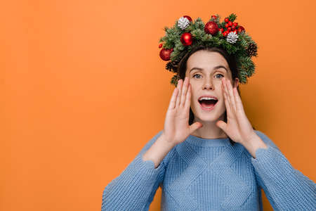 Cute young woman in traditional Christmas wreath, openes mouth wide, screams joyfully, Xmas is...