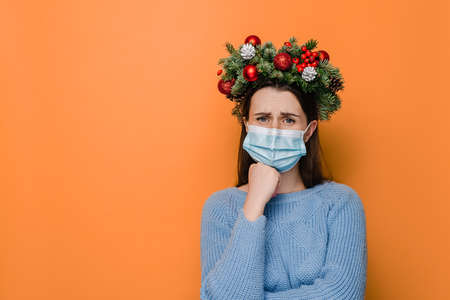 Grumpy displeased female in medical face mask, keeps hands half crossed, holds chin, has angry expression, being deep in thoughts, dressed in winter sweater, models over orange studio backgroundの写真素材