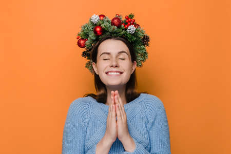 Cheerful woman in Christmas wreath hands folded in prayer beg about something good, dressed in winter blue sweater, make wish, isolated on orange studio background. Happy New Year merry holidayの写真素材