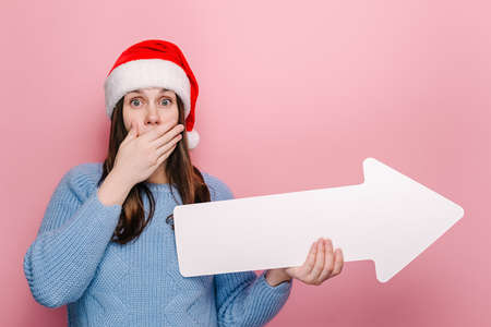 Shocked excited young woman holding white arrow, covering his mouth with his hand, wears cozy sweater and Christmas hat, isolated on pink background studio. Happy New Year celebration merry holidayの写真素材