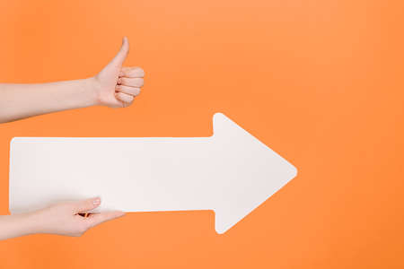 Close up of female hand holding big paper arrow and makes gesture thumb up gesture, demonstrates approval or agreement, isolated on pastel orange studio background with copy space for advertisementの写真素材