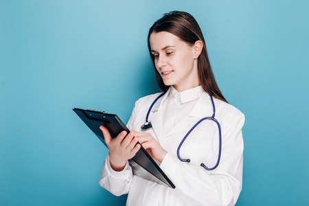 Portrait of cute professional young woman physician specialist in white coat standing on blue studio background with copy space, writing down symptoms. Healthcare workers and vaccination conceptの写真素材