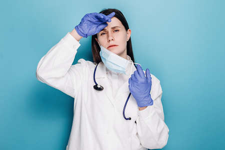 Young doctor woman or nurse in gloves and mask suffering from headache desperate and stressed because pain and migraine, wears stethoscope and white coat, isolated over blue background. Hand on headの写真素材