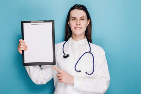 Professional cute woman doctor pointing at clipboard paper, explain disease or showing chart, wear white coat and stethoscope. Covid 19, preventing virus, healthcare workers and vaccination conceptの写真素材