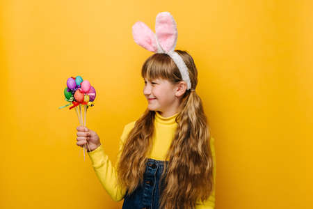 Portrait of cute happy school girl kid with bunny fluffy ears, holds and looking on small painted eggs on sticks, going to celebrate Easter, stands over yellow studio wall. Spring holiday conceptの写真素材