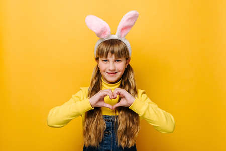 Smiling cute school girl in pink bunny fluffy ears showing heart gesture, isolated on yellow studio...