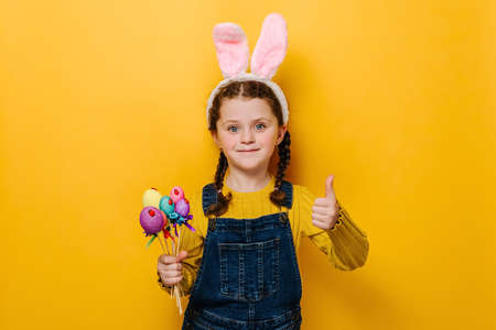 Charming little girl kid holding small colored eggs and showing thumb up as sign of approval or agreement isolated over yellow background, wears bunny fluffy ears. Acceptance okay and Easter conceptの写真素材