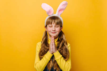 Portrait of cute beautiful teenager girl in pink bunny fluffy ears is praying for something, wears sweater, isolated on yellow studio background with copy space. Easter holiday and wish conceptの写真素材