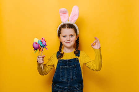 Lovely sweet preschool girl in pink bunny fluffy ears holding colored eggs on sticks and makes okay gesture, agrees with good proposal, isolated over yellow studio background. Happy Easter conceptの写真素材