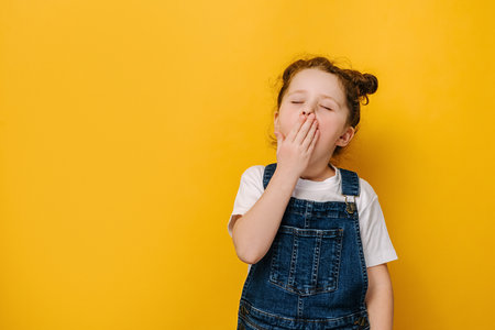 Little preschool girl feeling tired or sleepy cover mouth with hand closed eyes yawning, isolated on yellow studio background with copy space, face adorable preschool kid suffer. Boredom conceptの写真素材