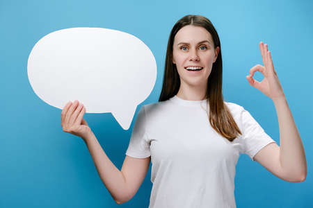 Portrait of cute beautiful young happy woman holding speech bubble posing isolated over blue studio background wall with copy space make okay gesture, dressed in white t-shirt. Hand sign conceptの写真素材