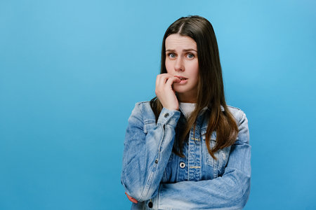 Worried young girl folded hands together near mouth, gnawing nails, young woman feels uncertain, stressed. Confused brunette female with frighten or panic gesture, isolated on blue studio backgroundの写真素材