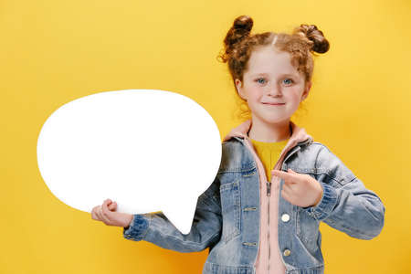 Portrait of cute little girl kid holding and pointing at blank speech bubble, wears denim jacket,...