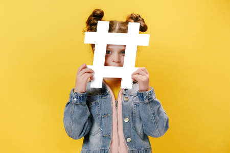 Joyful cute little girl holding hashtag symbol and looking fun at camera with toothy smile, showing hash sign, isolated on yellow studio background. Concept of trendy social media posts and bloggingの写真素材