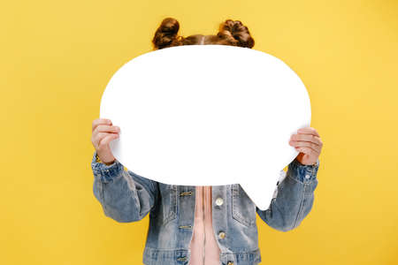 Close up of little girl child holding an big white empty speech bubble in front of his head, wears denim jacket, posing isolated on pastel yellow colored background in studio. Mock up copy spaceの写真素材