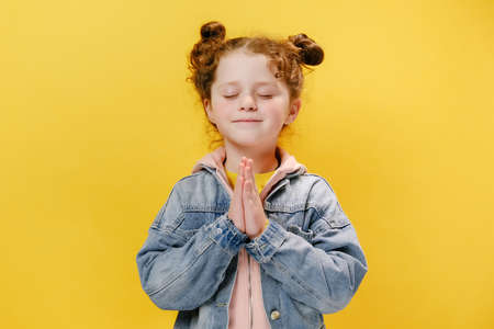Happy beautiful little girl child join hands pray with hope meditating, isolated on yellow studio background wall, calm religious small kid feel gratitude thanking in, faith meditation conceptの写真素材