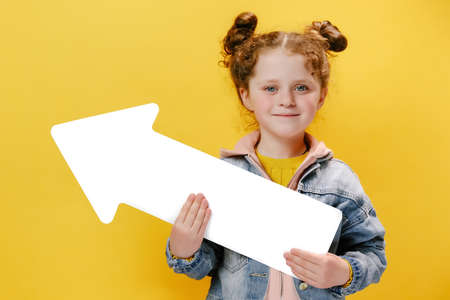 Cute smiling friendly little girl kid hold big paper arrow, looking happy at camera, wears denim jacket, isolated on yellow studio background with copy space for advertisement. People emotion conceptの写真素材