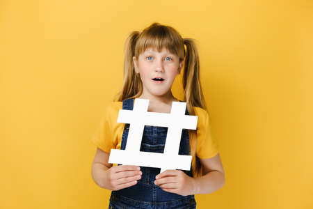 Funny excited surprised little girl kid holding big white hashtag symbol and looking with amazement,...