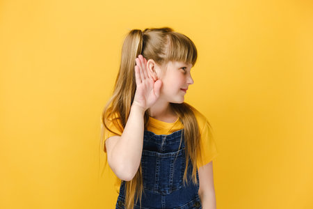 Curious little girl kid likes gossips, wants to overhear secret information, tries to hear conversation, listens attentively keeps hand near ear for eavesdropping, isolated on yellow studio backgroundの写真素材