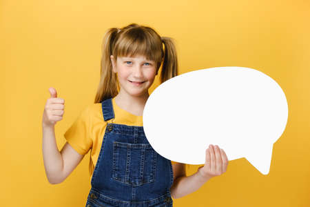 Positive nice cute girl child holding white empty blank Say cloud, speech bubble showing thumb up,...