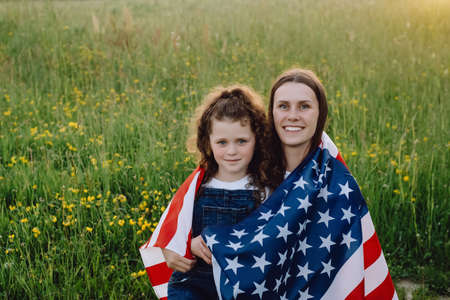 Portrait of attractive smiling young mother with cute daughter kid wrapped in american flag, happy...