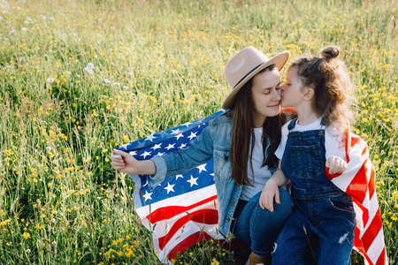 Patriotic cute young mother in hat and little daughter kid holds usa United States flag sitting on meadow during sunset. Smiling free proud independent family feeling freedom, independence conceptの写真素材
